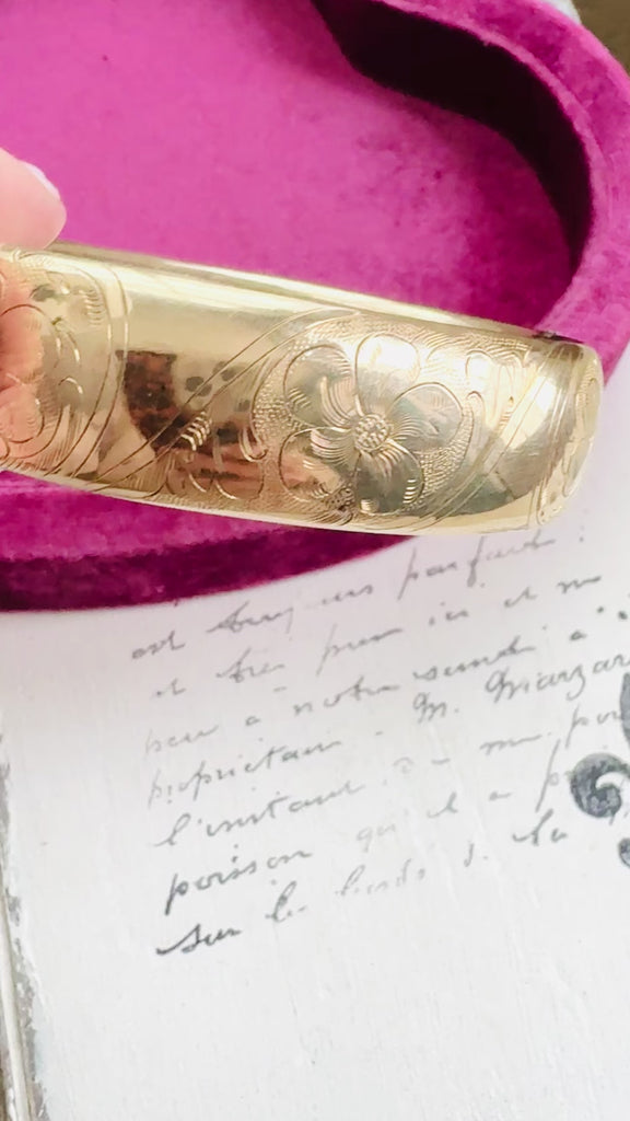 Hinged gold bangle bracelet styled on soft script background.