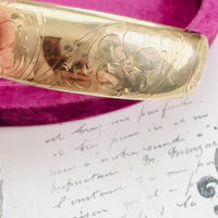 Hinged gold bangle bracelet styled on soft script background.